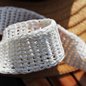 May include: White crocheted trim with a woven texture. The trim is folded and shows the intricate detail of the crocheted stitches.
