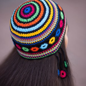 May include: A colorful crocheted hat with a circular design and tassels. The hat is made with a variety of bright colors, including red, orange, yellow, green, blue, and black.