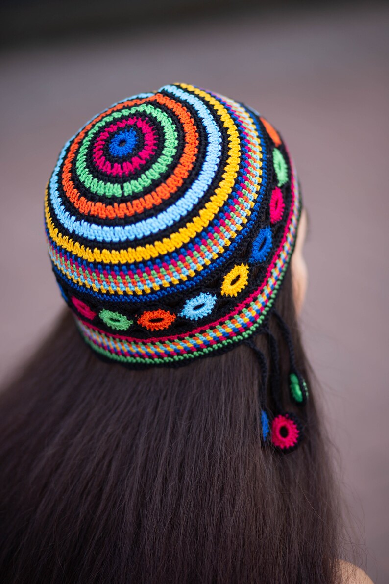 May include: A colorful crocheted hat with a circular design and tassels. The hat is made with a variety of bright colors, including red, orange, yellow, green, blue, and black.