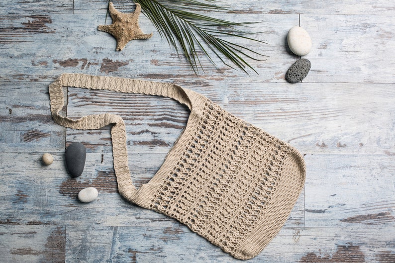 May include: A beige crocheted tote bag with a long strap, laying on a light blue wooden surface with a starfish, palm leaves, and stones.