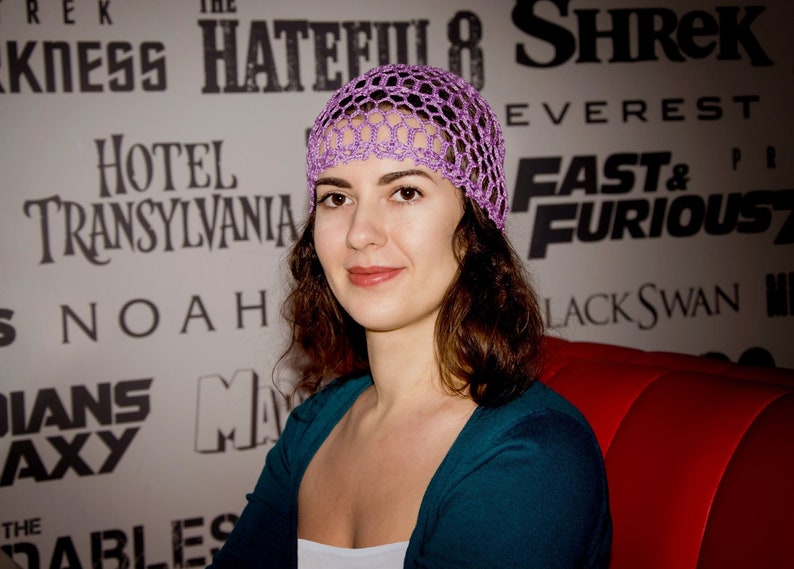 May include: A woman with long brown hair wearing a purple crochet hat and a teal blue sweater. She is sitting on a red couch in front of a wall with movie posters.
