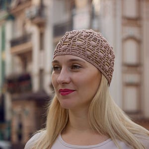 May include: A woman with blonde hair is wearing a light brown crocheted hat. The hat has a delicate openwork design. She is looking to the side and smiling.