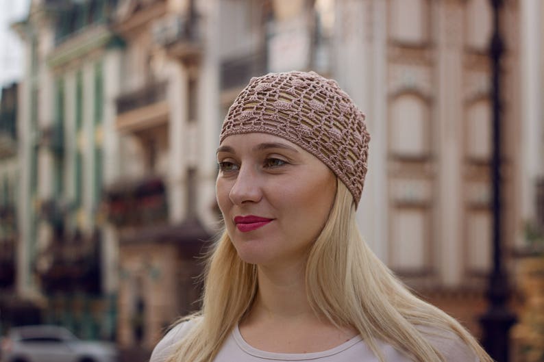 May include: A woman with blonde hair is wearing a light brown crocheted hat. The hat has a delicate openwork design. She is looking to the side and smiling.