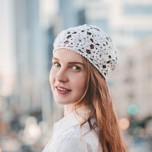 A young woman stands in a half-turn wearing a floral French beret, crocheted with motifs. A woman wearing white flowers summer hat.