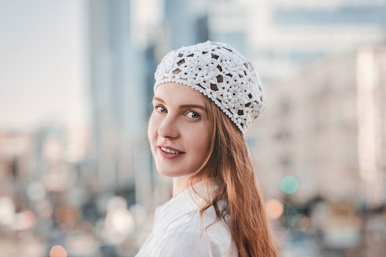 A young woman stands in a half-turn wearing a floral French beret, crocheted with motifs. A woman wearing white flowers summer hat.