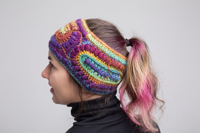 May include: A colorful crocheted headband with a multi-colored pattern. The headband is worn by a person with a ponytail.