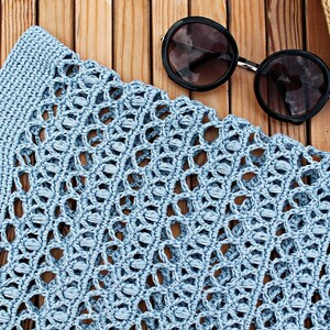 May include: A light blue crocheted tote bag with a woven pattern. The bag is lying on a wooden surface with a pair of round sunglasses and a straw hat.