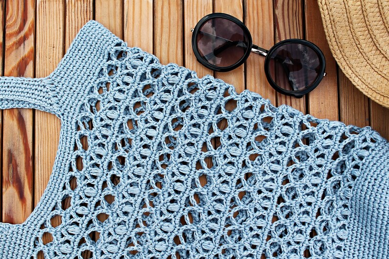 May include: A light blue crocheted tote bag with a woven pattern. The bag is lying on a wooden surface with a pair of round sunglasses and a straw hat.