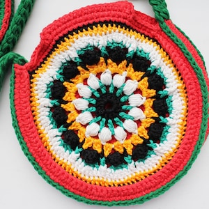 May include: A round crocheted bag with a colorful mandala design in red, yellow, green, white, and black. The bag has a green strap.