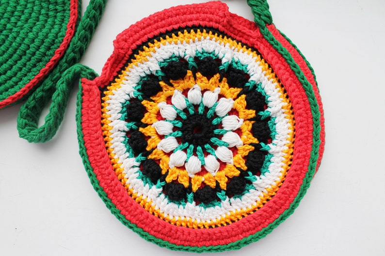 May include: A round crocheted bag with a colorful mandala design in red, yellow, green, white, and black. The bag has a green strap.