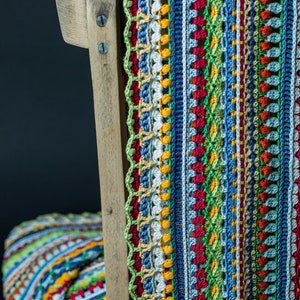 A crocheted, bright, multi-colored scarf with an intricate pattern is hanging on a chair, set against a black background.