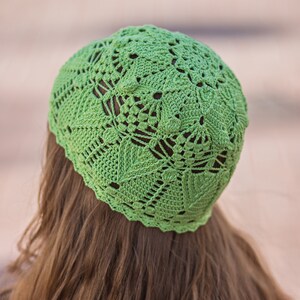 May include: A green crocheted hat with a lacy design. The hat is worn on a person with long brown hair.