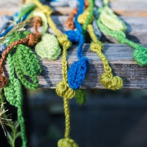 May include: A crocheted garland with green, blue, and brown leaves. The garland is made of yarn and is hanging from a wooden surface.