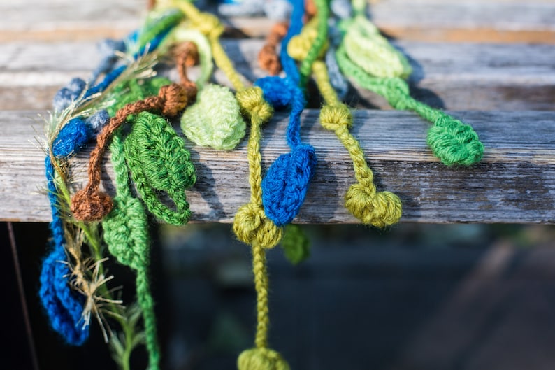 May include: A crocheted garland with green, blue, and brown leaves. The garland is made of yarn and is hanging from a wooden surface.