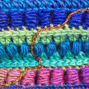 May include: Close-up of a multi-colored crocheted fabric with a gold chain woven into the design. The fabric is made of different shades of blue, green, pink, and purple yarn.