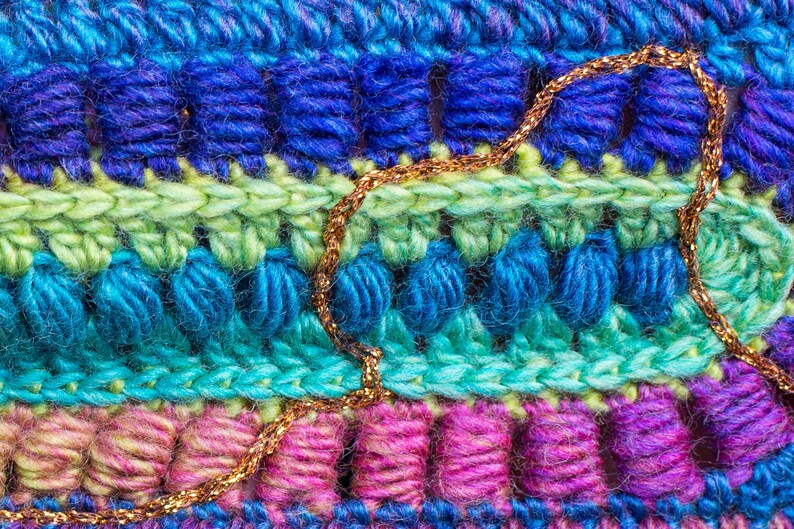May include: Close-up of a multi-colored crocheted fabric with a gold chain woven into the design. The fabric is made of different shades of blue, green, pink, and purple yarn.