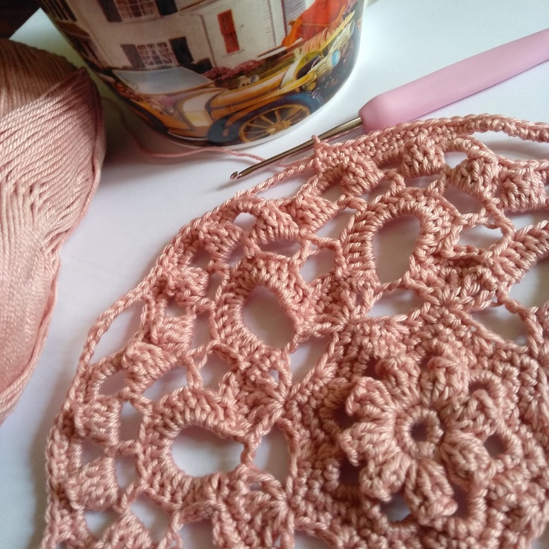 May include: A close-up of a pink crocheted doily with a floral design. The doily is being worked on with a pink crochet hook.