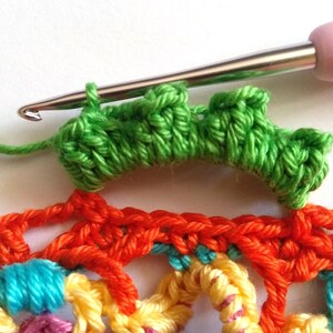 May include: A close-up of a crochet project with a green crocheted row on top of a multi-colored crocheted piece. A crochet hook is visible in the image.