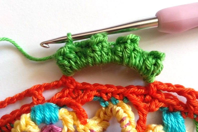 May include: A close-up of a crochet project with a green crocheted row on top of a multi-colored crocheted piece. A crochet hook is visible in the image.