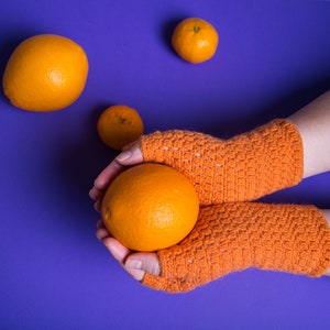 Könnte beinhalten: Ein Paar orangefarbene Fingerlose Handschuhe aus Häkelarbeit auf einem violetten Hintergrund, mit einer Person, die eine Orange in ihren Händen hält.