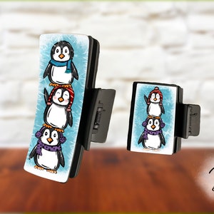 May include: Three penguins wearing winter hats and scarves are stacked on top of each other on a blue background with a white border. The penguins are on a black phone grip.