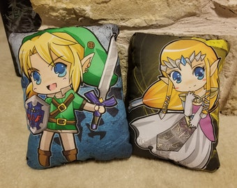 May include: Two plush pillows featuring cartoon characters from the Legend of Zelda video game series. The pillow on the left features Link, a young boy with blonde hair, wearing a green tunic and holding a sword. The pillow on the right features Zelda, a young girl with blonde hair, wearing a white dress and holding a sword.