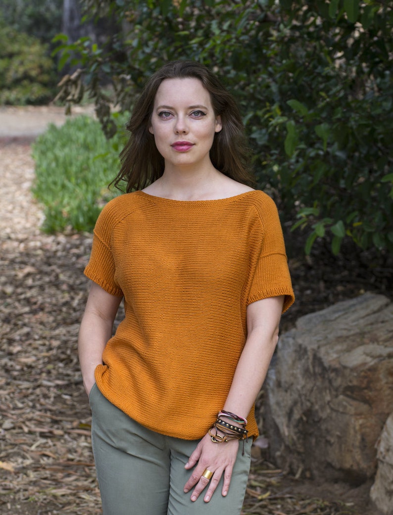 May include: A woman wearing a burnt orange knitted sweater with short sleeves and green pants. She is wearing several bracelets on her wrist.