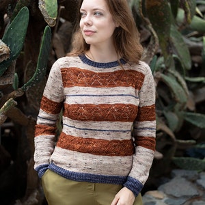 May include: A woman wearing a striped sweater with a brown, blue, and white color scheme. The sweater has a cable knit pattern and is worn with olive green pants.