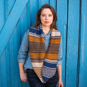May include: A woman wearing a blue denim shirt and jeans stands in front of a blue wooden wall. She is wearing a colorful striped infinity scarf with a geometric pattern.