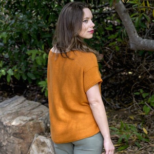 May include: A woman wearing a rust-colored knitted top with short sleeves and light green pants. She is standing in a wooded area with green foliage and a tree trunk in the background.