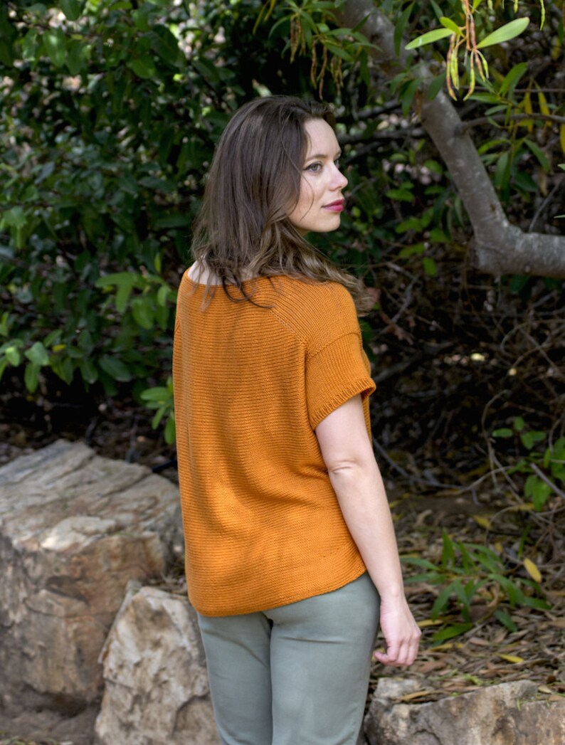 May include: A woman wearing a rust-colored knitted top with short sleeves and light green pants. She is standing in a wooded area with green foliage and a tree trunk in the background.