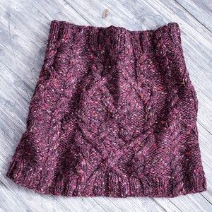 Puede incluir: Una falda de punto de color burdeos oscuro con un patrón de punto de cable. La falda tiene una textura moteada con motas de marrón, verde y rojo.