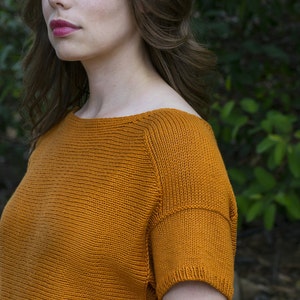 May include: A close-up of a woman wearing a burnt orange knitted top with a boat neck and short sleeves.