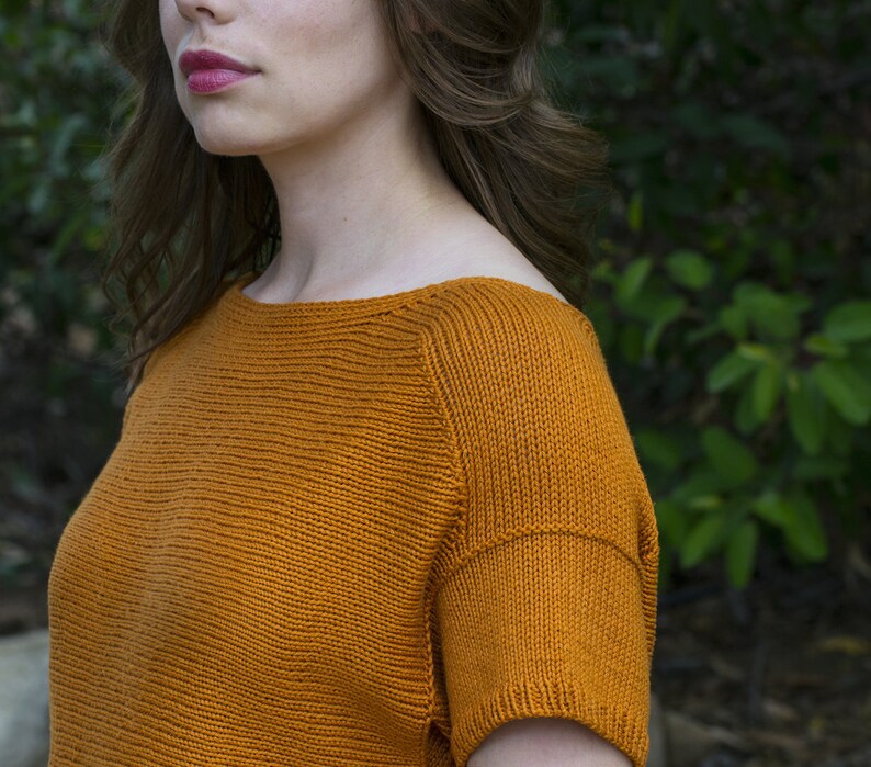 May include: A close-up of a woman wearing a burnt orange knitted top with a boat neck and short sleeves.