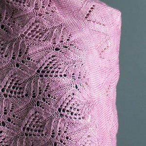 May include: A pink knitted shawl with a delicate lace pattern. The shawl is draped over a mannequin and has a soft, flowing texture.