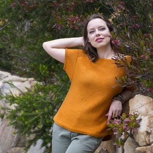 May include: A woman wearing a burnt orange knitted top with short sleeves and a relaxed fit. She is standing in front of a rock wall with green foliage.