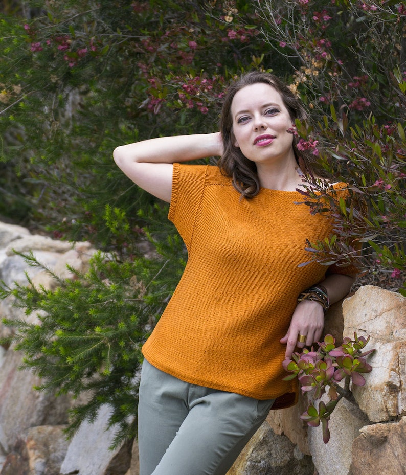May include: A woman wearing a burnt orange knitted top with short sleeves and a relaxed fit. She is standing in front of a rock wall with green foliage.