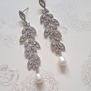 May include: A pair of long, silver earrings with a delicate, leaf-shaped design. Each earring features a cluster of sparkling clear stones and a single, white pearl drop.