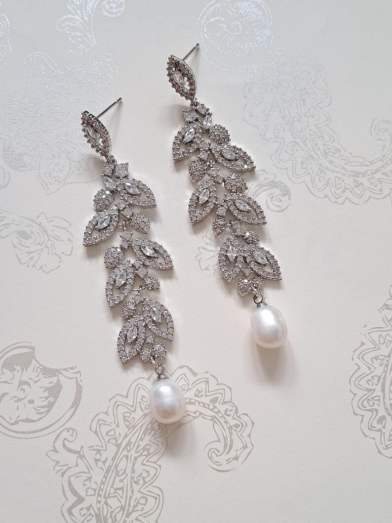 May include: A pair of long, silver earrings with a delicate, leaf-shaped design. Each earring features a cluster of sparkling clear stones and a single, white pearl drop.