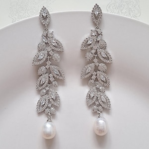 May include: A pair of silver dangle earrings with a pearl drop. The earrings feature a design of multiple leaves with clear cubic zirconia stones.