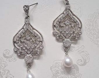 Könnte beinhalten: Ein Paar silberfarbene Chandelier-Ohrringe. Jeder Ohrring hat ein verziertes, offenes Design, das mit klaren Kristallen besetzt ist. Ein großer, runder Kristall und eine tropfenförmige Perle hängen an jedem Ohrring und erzeugen einen glamourösen Look.