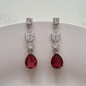 May include: A pair of silver dangle earrings featuring a teardrop-shaped red gemstone and clear cubic zirconia accents.