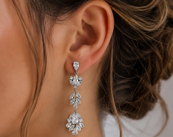 May include: A silver-toned drop earring featuring a vertical arrangement of clear, faceted stones. The design includes a single pear-shaped stone at the top, followed by a marquise-shaped stone, a leaf-like cluster, and a larger, ornate floral-shaped base.