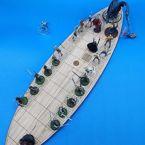 May include: A wooden game board shaped like a ship with miniature figures of people and a dragon. The figures are painted in various colors and are arranged on the board in a battle scene. The board is made of light wood and has a grid pattern.