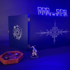 May include: A dark blue wooden game box with intricate white geometric designs. The box is open, revealing several game cards with text. A small figurine and a dice tray with dice are in front of the box.