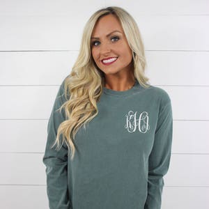 May include: A long-sleeved, teal-colored t-shirt with a white monogram on the left chest. The shirt is made of a soft, comfortable fabric and has a relaxed fit.