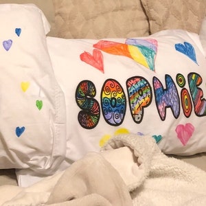 May include: Two white pillowcases with colorful drawings on them. One pillowcase has the word "Josh" written in rainbow colors. The other pillowcase has the word "Sophie" written in rainbow colors with swirls and other designs.