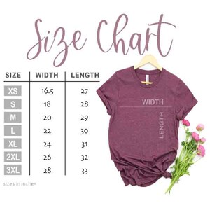 Puede incluir: Tabla de tallas para una camiseta con medidas en pulgadas para el ancho y la longitud. Las tallas incluyen XS, S, M, L, XL, 2XL y 3XL. Se muestra una camiseta granate sobre un fondo blanco.