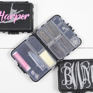 May include: Three black plastic containers with clear lids. The containers are small and rectangular. One container has the word "Harper" written on it in pink and gray letters. Another container has a monogram "BVM" written on it in silver letters. The third container is open and has various hair accessories inside.