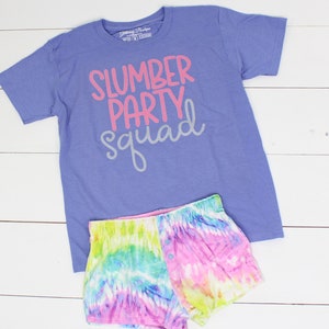 Może przedstawiać: Fioletowy T-shirt z napisem "slumber party squad" w r&oacute;żowych i szarych literach. T-shirt jest połączony z kr&oacute;tkimi spodenkami tie-dye w odcieniach r&oacute;żowego, niebieskiego, zielonego i ż&oacute;łtego.